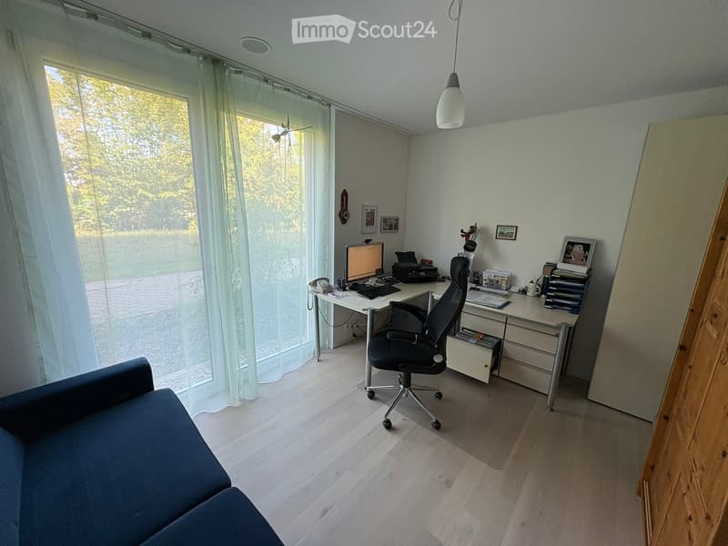Bright room with wooden flooring, large window with sheer curtains, desk with computer and chair, sofa, and wall decorations.