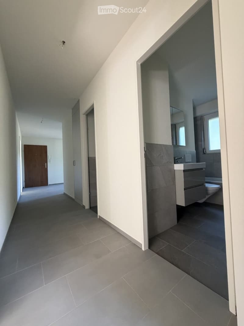 Modern hallway with grey tiled flooring, white walls, and a partially visible bathroom with a sink and toilet.