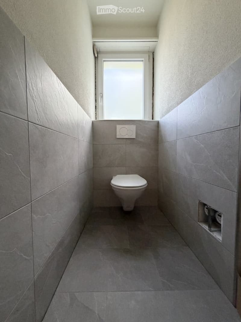 Modern, minimalist bathroom with grey tiles, a toilet, and a small window above it.