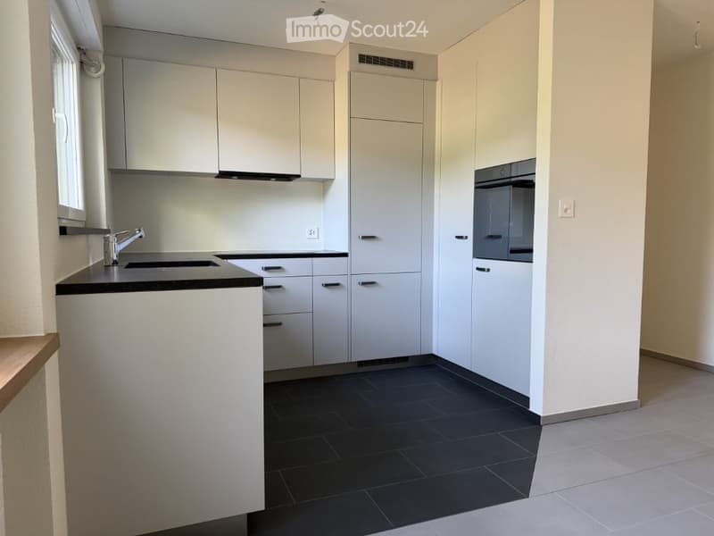 Modern kitchen with white cabinets, black countertop, stainless steel sink, and built-in oven. Dark tiled floor and light-colored walls.