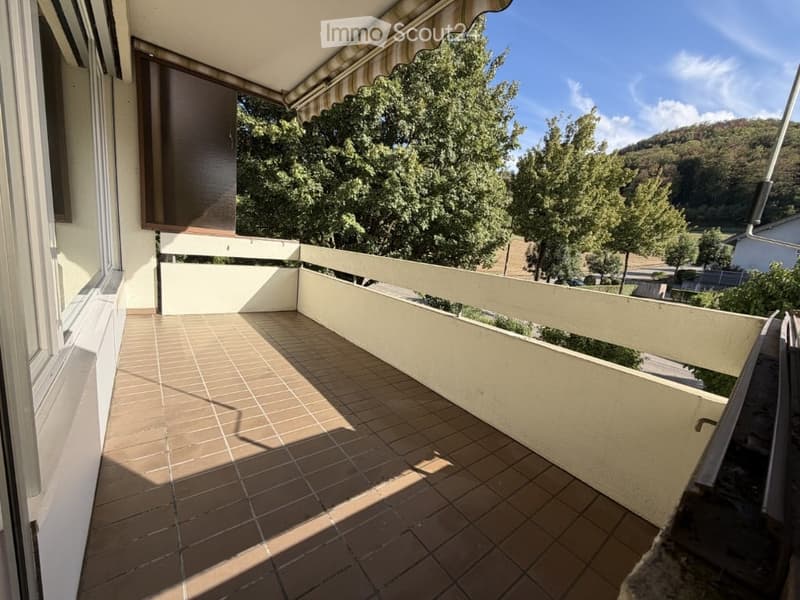 Balcony with tiled floor, white railing, and striped awning, overlooking a green landscape with trees and hills.