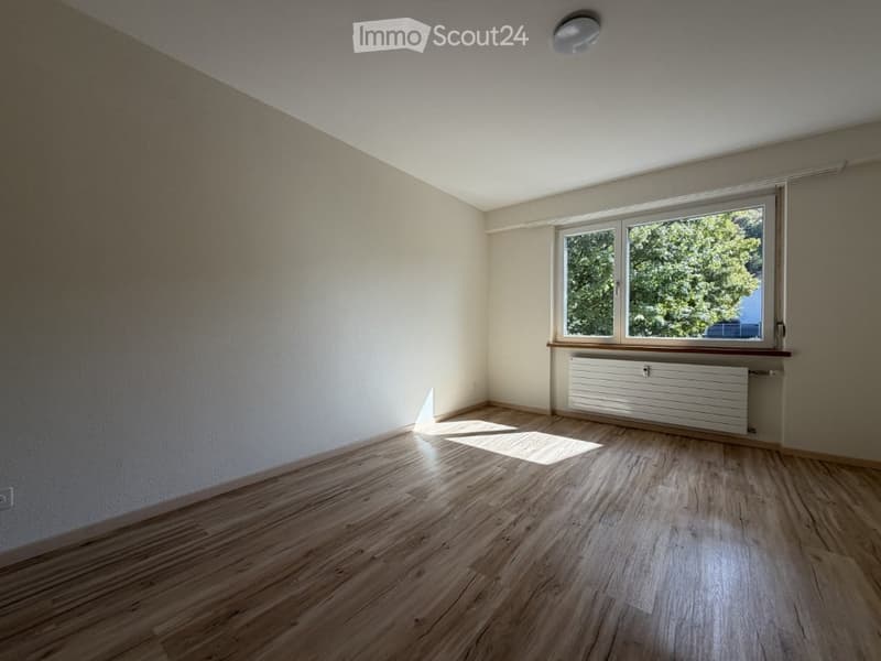 Empty room with wooden flooring, white walls, and a large window with a view of greenery outside. A radiator is visible beneath the window.