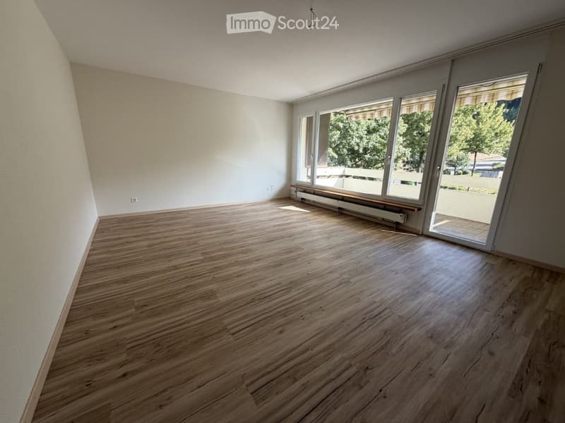 Empty room with light wooden flooring, white walls, large windows, and a sliding glass door leading to a balcony.