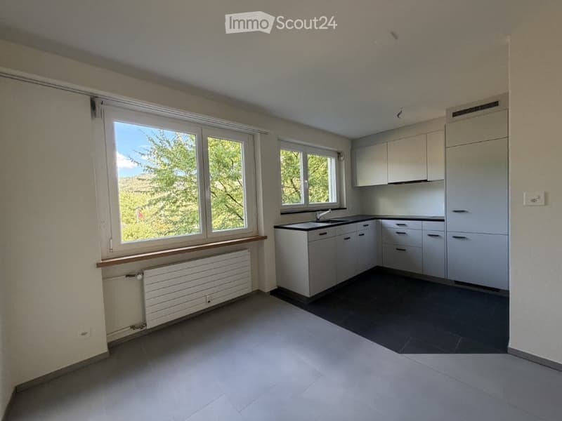 Modern kitchen with white cabinets, dark countertops, and large windows offering a view of greenery outside.