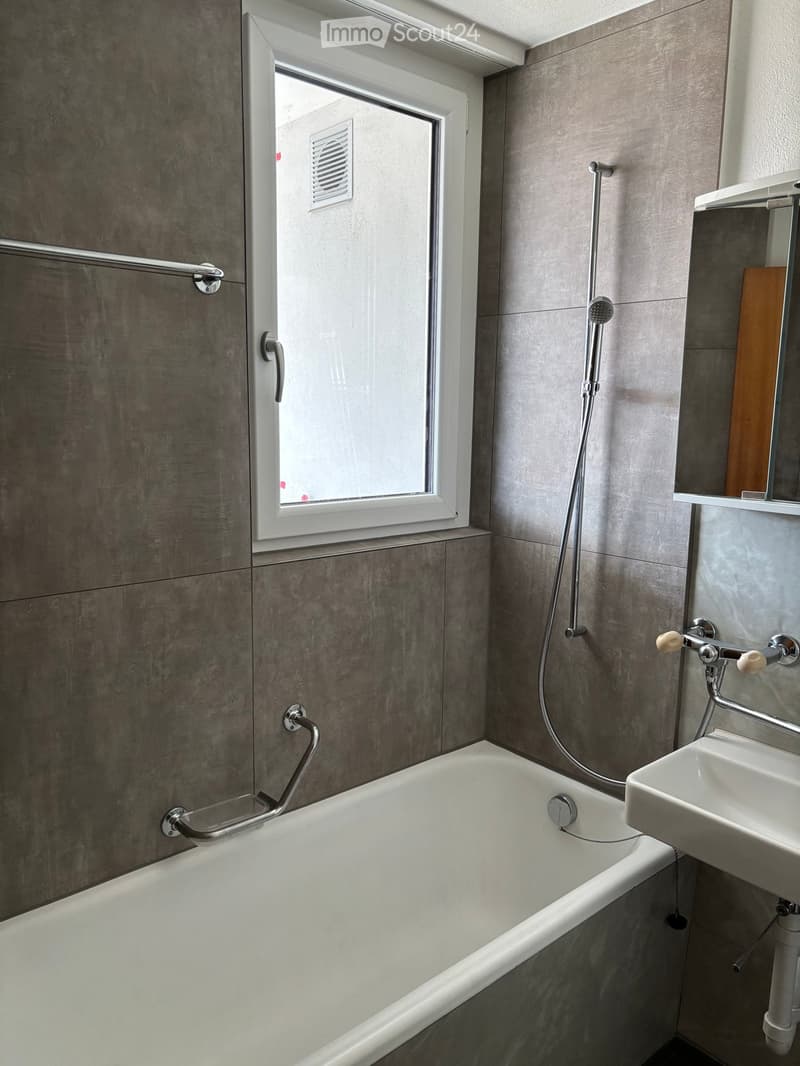Modern bathroom with a bathtub, sink, showerhead, and handrail. Grey tiled walls and a window provide natural light.
