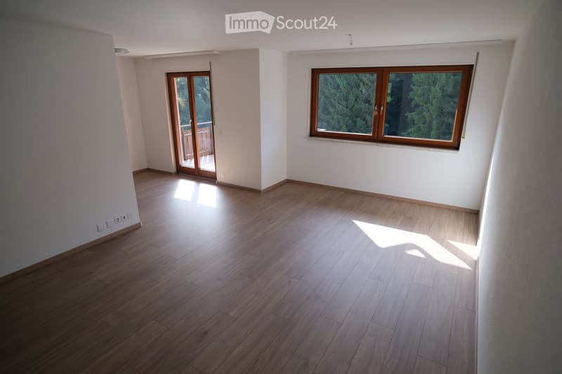 Empty room with wooden flooring, white walls, and large windows with wooden frames. Features a sliding glass door leading to a balcony.