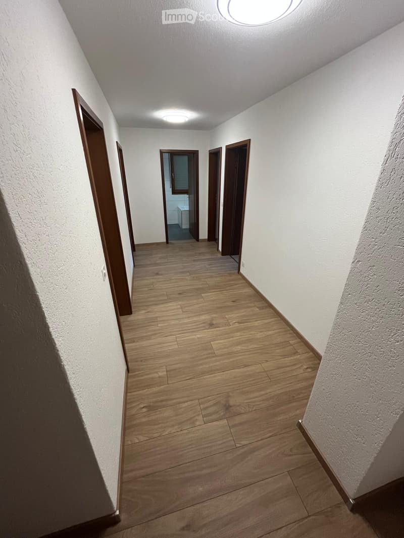 A clean, well-lit hallway with wooden flooring and white walls, featuring several closed doors and recessed ceiling lights.