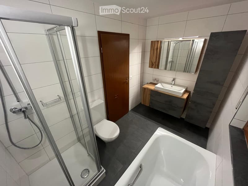 Modern bathroom with a bathtub, toilet, sink, mirror, and a glass-enclosed shower. The space features white tiles, a wooden vanity, and a sleek design.