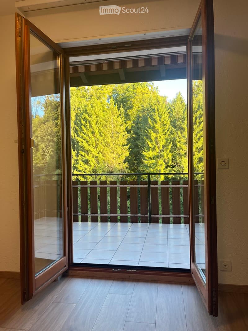 Open sliding glass doors lead to a tiled balcony with wooden railings, overlooking a lush green forest.