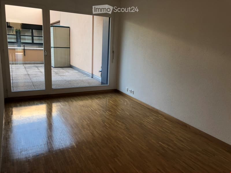 Empty room with wooden flooring, large sliding glass doors leading to a balcony, and a visible refrigerator in the adjacent space.