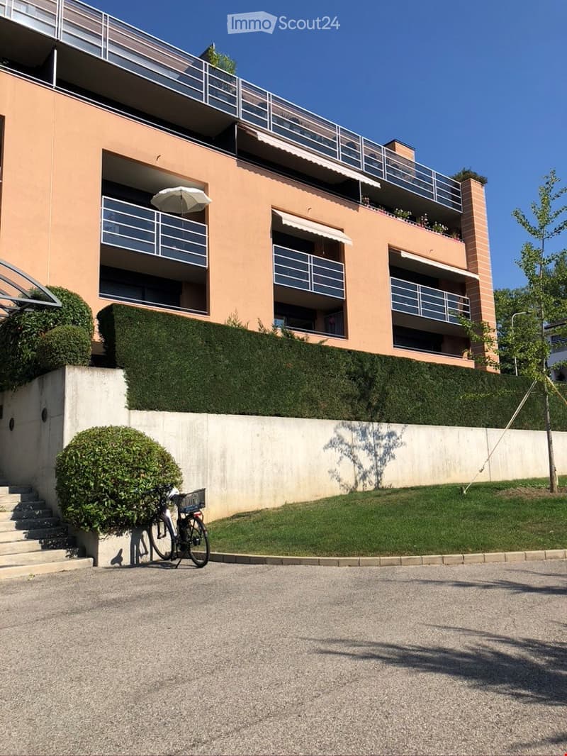 Modern multi-story building with balconies, light pink facade, and neatly trimmed hedges. A bicycle is parked near the entrance.