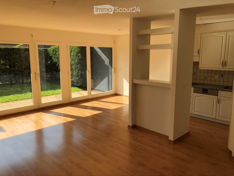 Bright, unfurnished living space with large glass doors leading to a garden, wooden flooring, and an adjacent kitchen area with white cabinets and a countertop.
