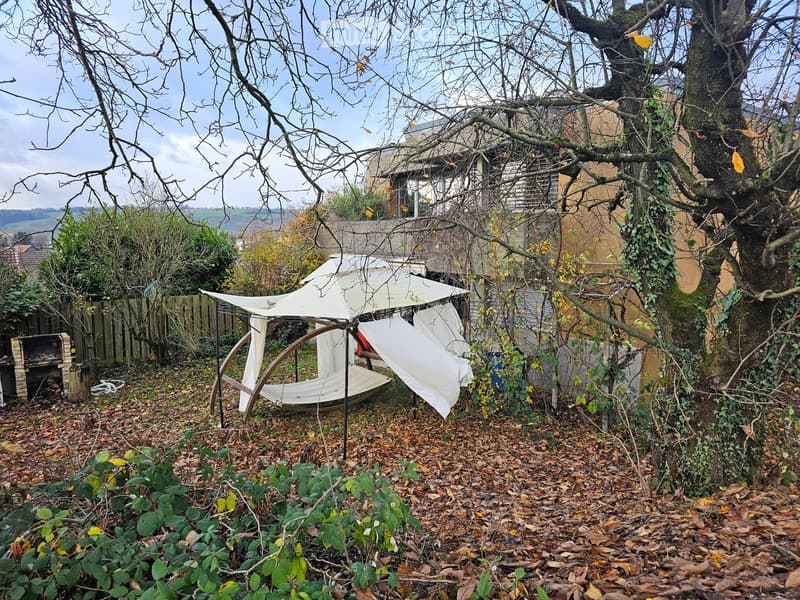 Garden with a wooden canopy, fallen leaves, tree, house wall in the background, fence, and small shed.
