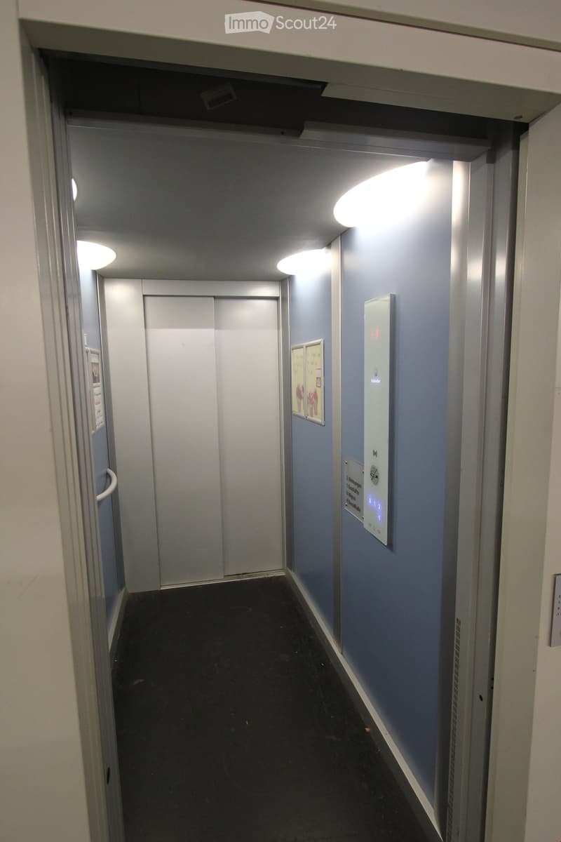 A modern hallway with light blue walls, black tiled floor, and illuminated ceiling lights. An elevator door is visible at the end of the corridor, along with a control panel and framed artwork on the walls.