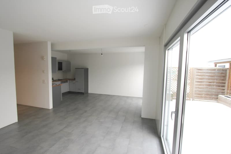 Open-plan living and dining area with grey tiled flooring, white walls, and a modern kitchen area featuring grey cabinets and a countertop. Large sliding glass doors lead to an outdoor space.