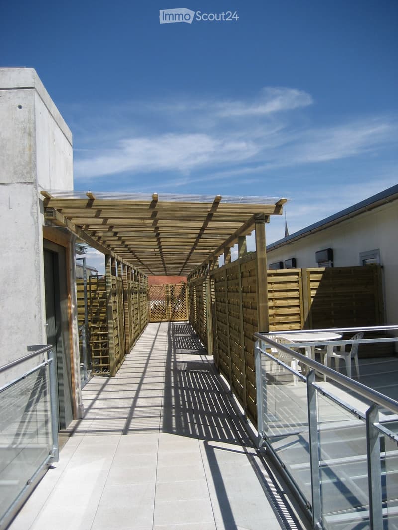 A covered outdoor walkway with wooden pergola, glass railings, and a small seating area with white plastic chairs and a table.