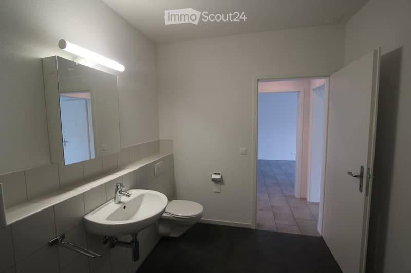 Modern bathroom with a white sink, toilet, mirror, and cabinet. The room has tiled walls and a dark floor, with an open doorway leading to another room.
