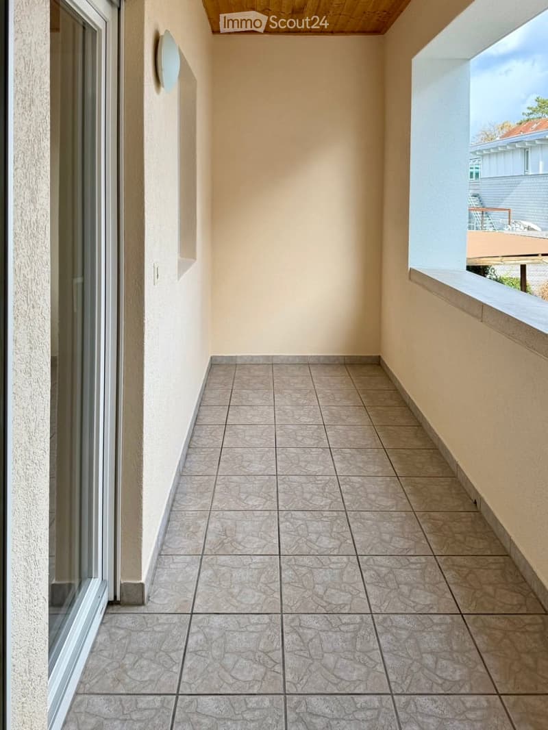 Balcony with tiled floor, beige walls, and wooden ceiling. Features large glass sliding doors and a view of neighboring buildings.