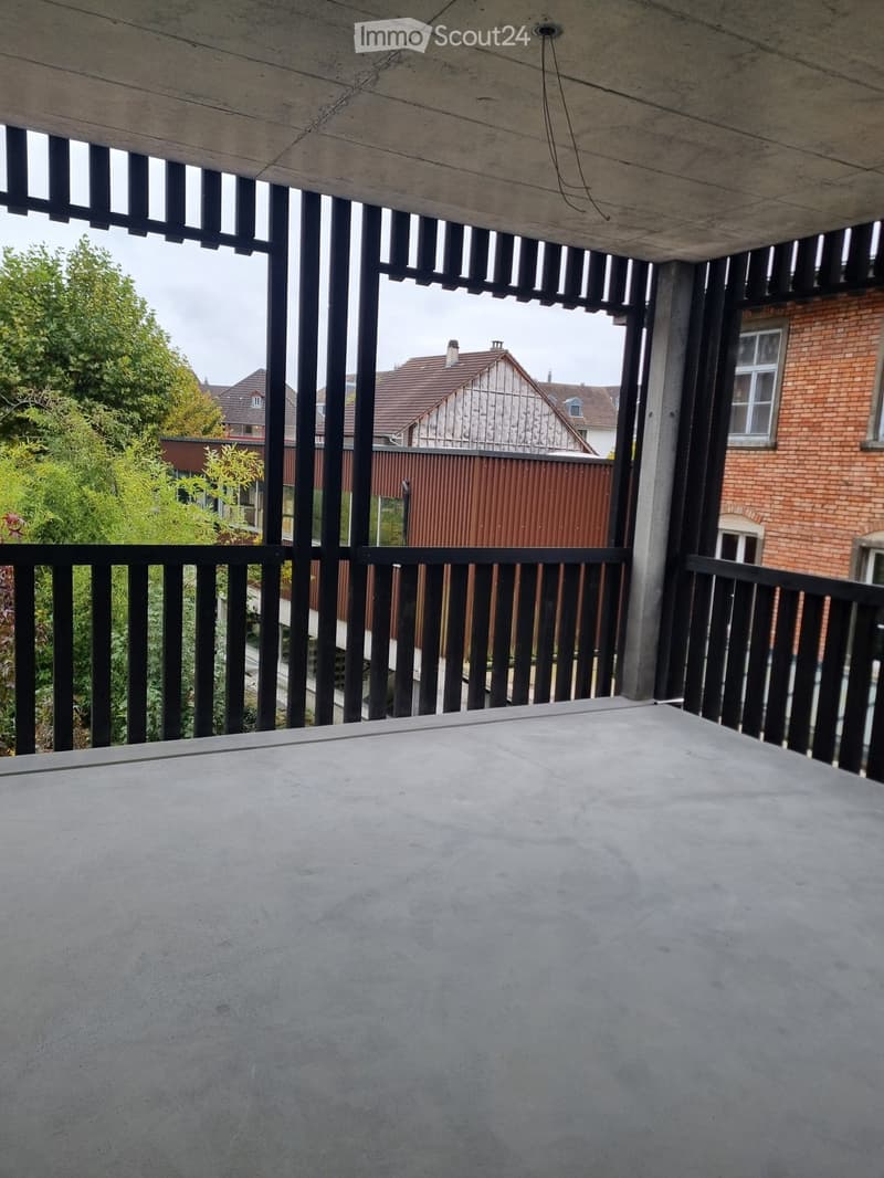 concrete floor, balcony with black railings, visible trees and building in the background