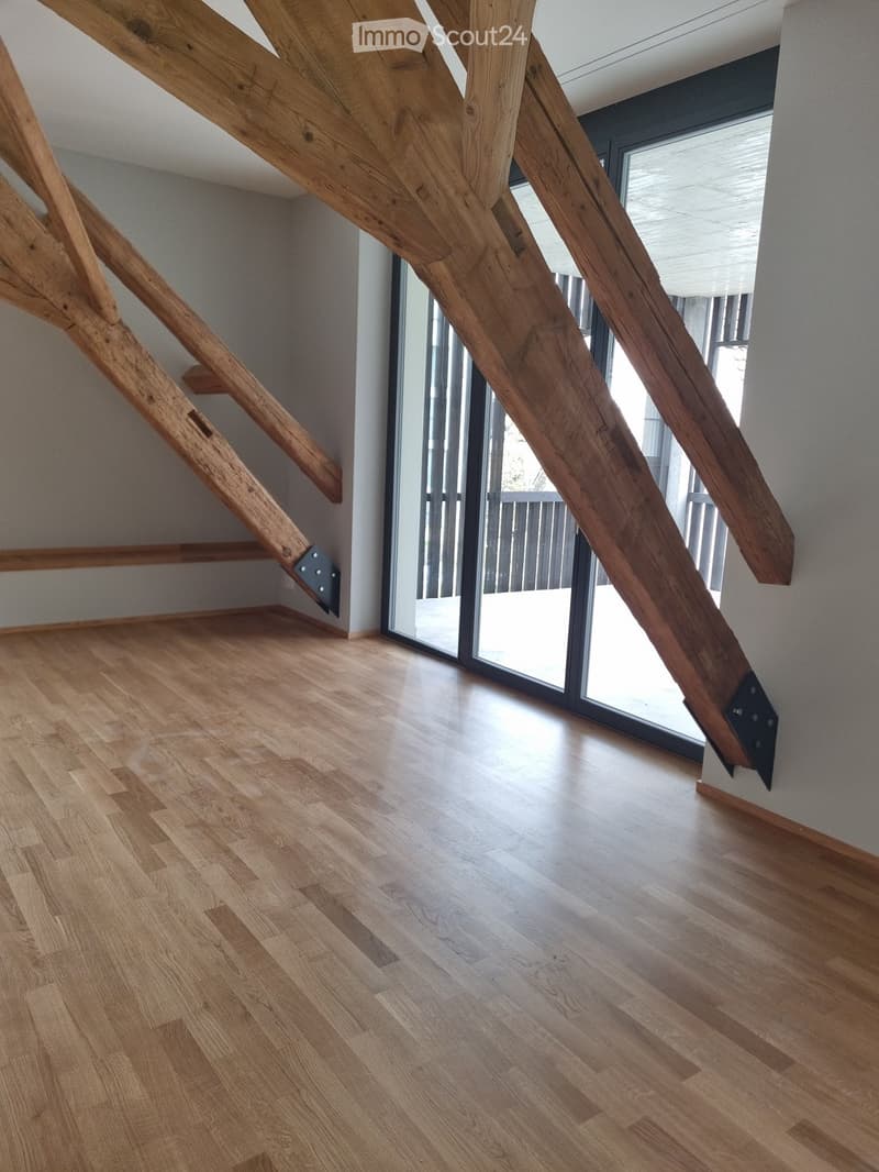 An attic with hardwood floors, exposed wooden trusses, sliding glass doors leading to balcony, white walls.