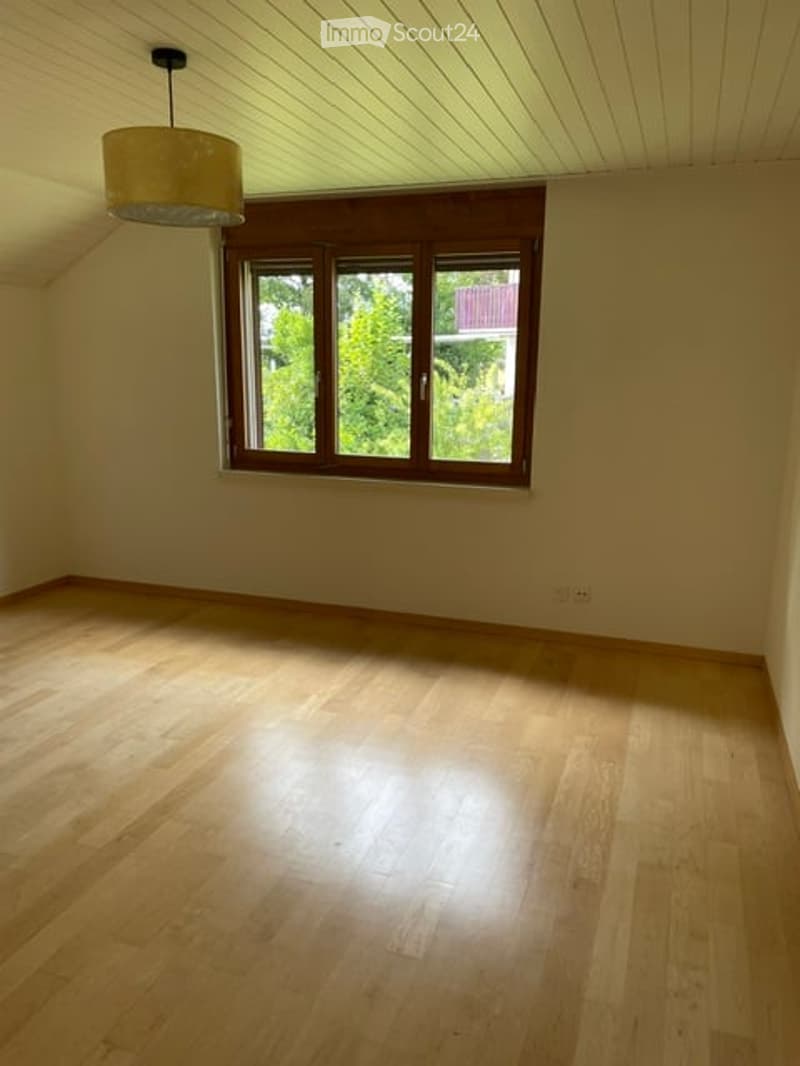 Empty room with wooden flooring, white walls, and a large window with wooden frame. A simple pendant light hangs from the ceiling.