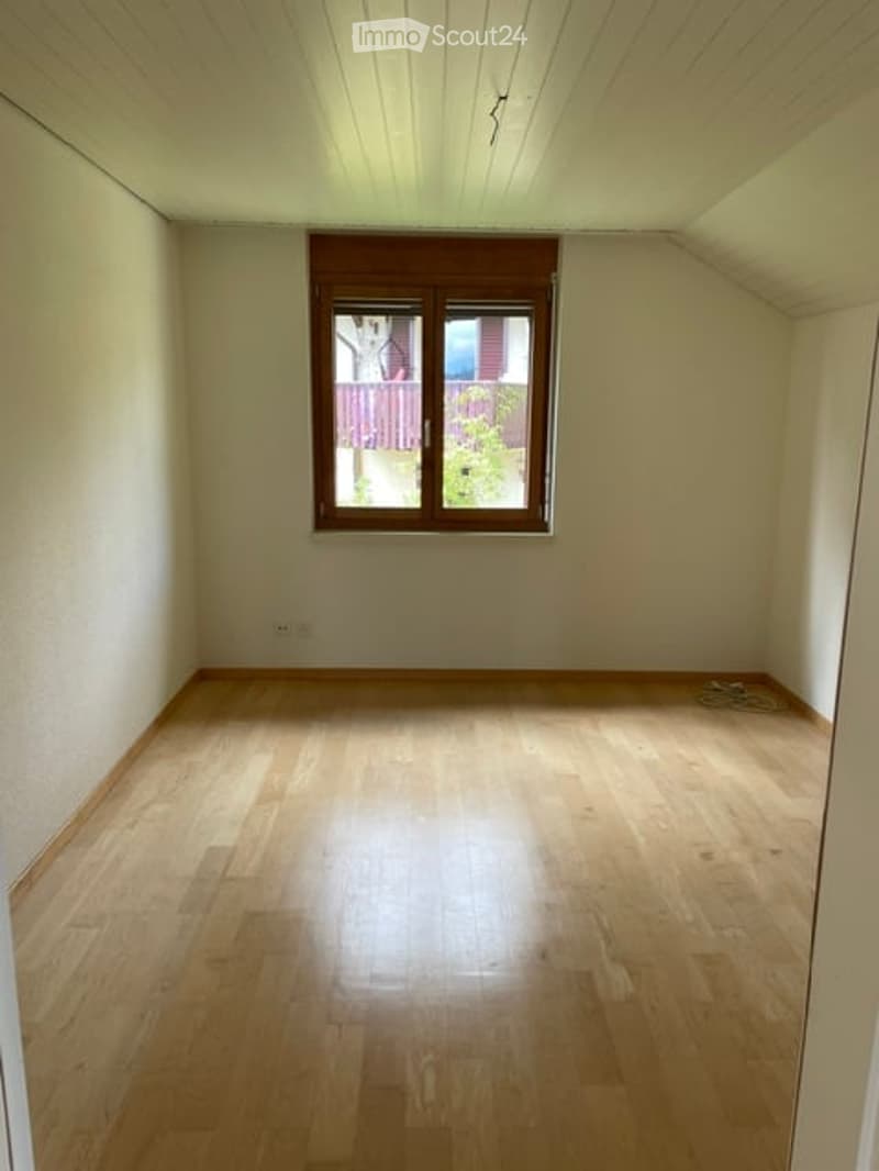 Empty room with wooden flooring and white walls, featuring a single window with wooden frame and a view of a garden.