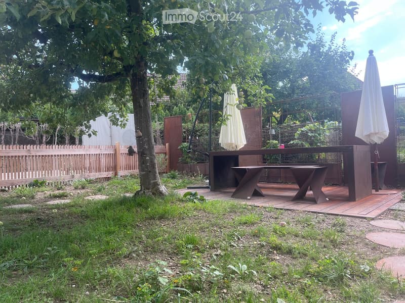 Garden with a wooden fence, a tree, a patio area with a table and chairs, and outdoor umbrellas.