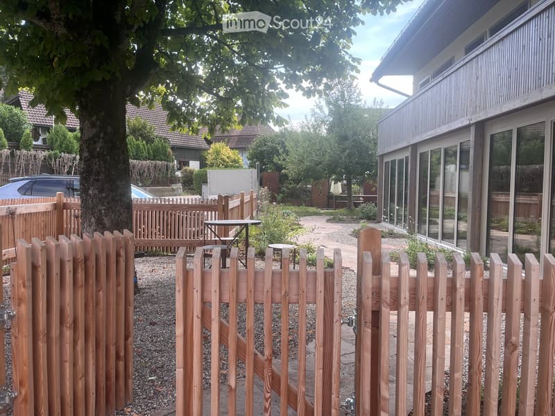 A well-maintained garden area enclosed by a wooden fence, featuring a tree, a small table, and a modern house with large windows in the background.