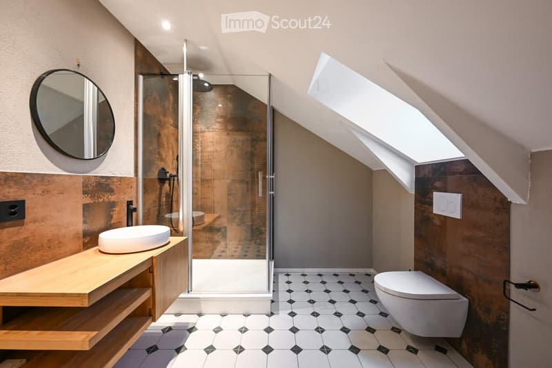Modern bathroom with a sleek, contemporary design. Features include a white sink on a wooden vanity, a toilet, a round wall-mounted mirror, and a glass-enclosed shower with a modern showerhead. The space has a sloped ceiling with skylights, black and whit