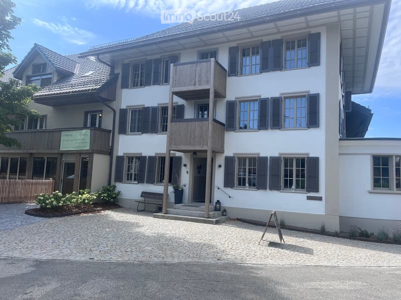 A two-story white building with multiple windows, shutters, and a balcony. The entrance features a small porch with steps, a bench, and potted plants. The building has a sign indicating it is available for rent.