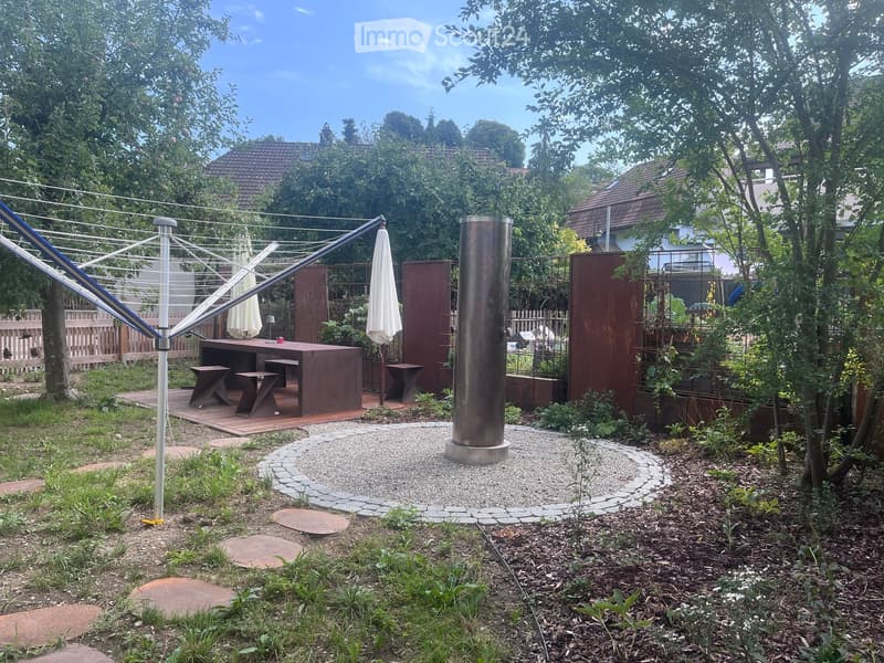 Garden with a wooden deck, outdoor dining table and chairs, umbrella, and a modern metal sculpture. The area is surrounded by trees and has a gravel pathway.