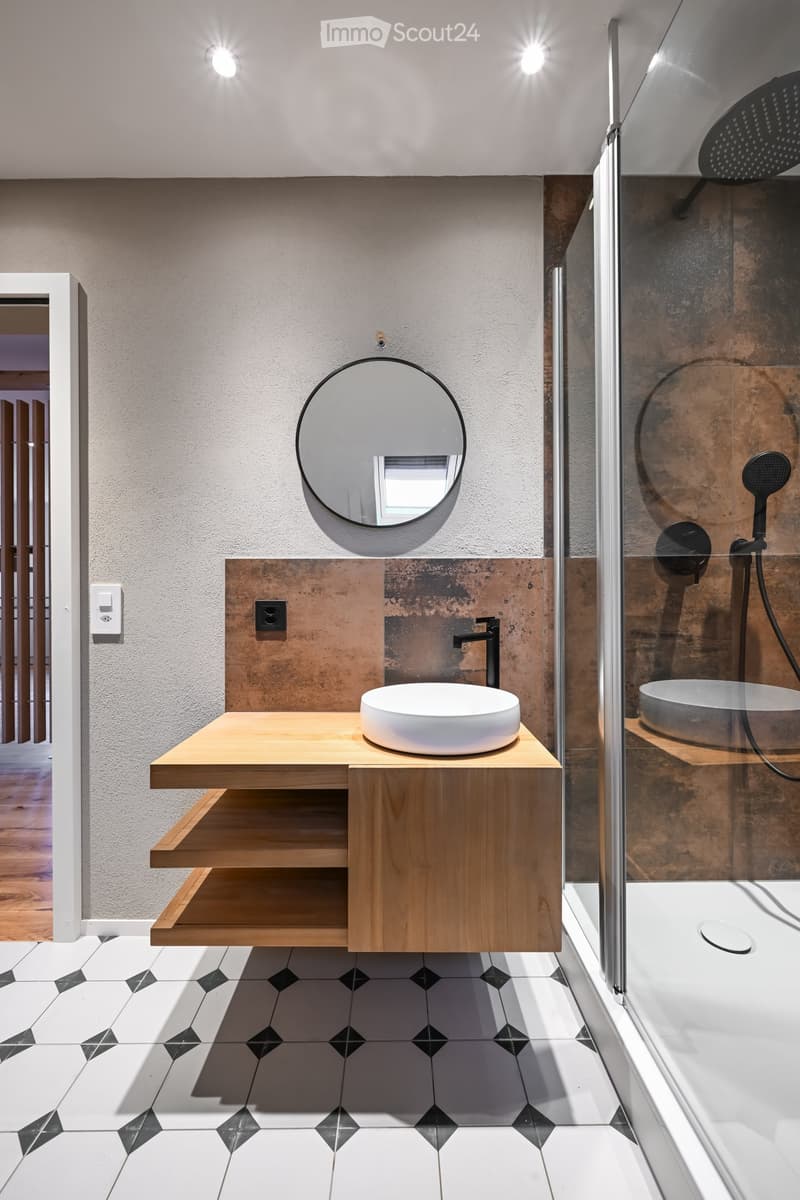 Modern bathroom with a wooden floating vanity, white sink, round mirror, and glass-enclosed shower with a black showerhead.