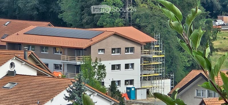 A multi-story residential building with red-tiled roofs, solar panels on the roof, and a partially constructed section with scaffolding.