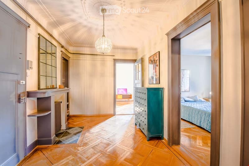 Elegant hallway with wooden flooring, ornate ceiling light, and open doorways leading to a living area and bedroom.