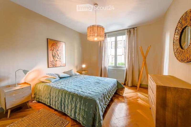 Cozy bedroom with a large bed, nightstands, a hanging woven lamp, a round mirror, and a painting on the wall. The room features wooden flooring and a small woven rug.