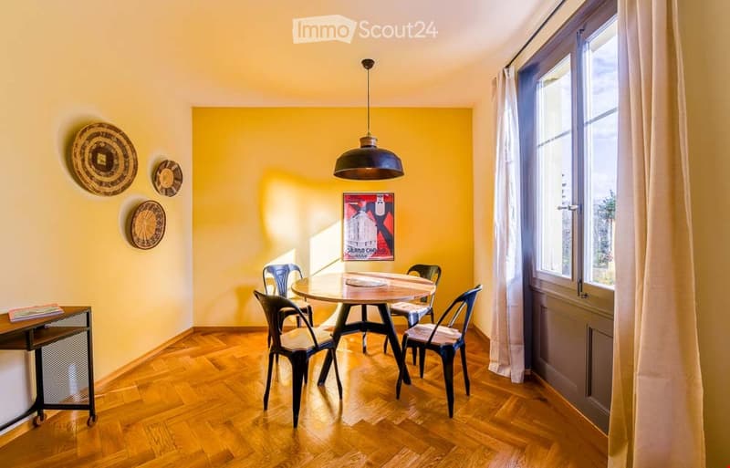 Bright dining area with a round wooden table surrounded by six black metal chairs, pendant light fixture, and wall art. Wooden floor and large window with white curtain.