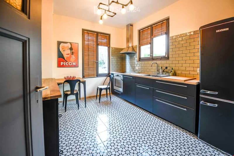 Modern kitchen with dark cabinets, stainless steel appliances, and patterned tile flooring. Features a pendant light fixture, a small dining area with chairs and a table, and a poster on the wall.