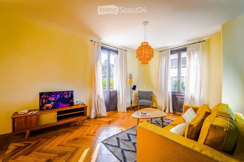 Bright living room with wooden flooring, large windows with white curtains, a yellow sofa, a coffee table, a television on a wooden stand, and a patterned rug.