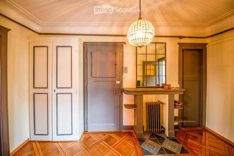 Elegant hallway with wooden herringbone flooring, ornate ceiling light fixture, large mirror, and built-in console table with radiator below.