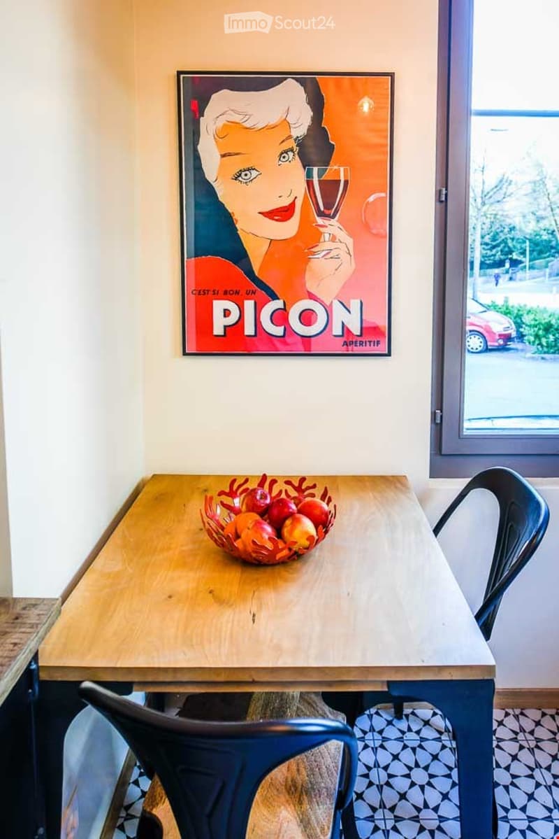 A small, modern dining area with a wooden table, two blue chairs, a decorative bowl of fruit, and a vintage-style poster on the wall.