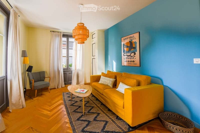 Bright living room with wooden flooring, blue accent wall, yellow sofa, coffee table, armchair, and decorative items.