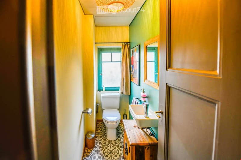 Cozy bathroom with yellow and green walls, a white toilet, a sink with a wooden vanity, a mirror, and a window with a curtain. The floor features a patterned tile design.