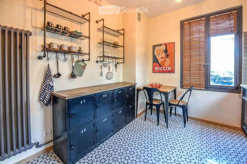A compact kitchen area with a dark blue cabinet, a wooden countertop, metal shelves, and a small dining table with two chairs. The floor features a patterned tile design, and a window with blinds is visible.