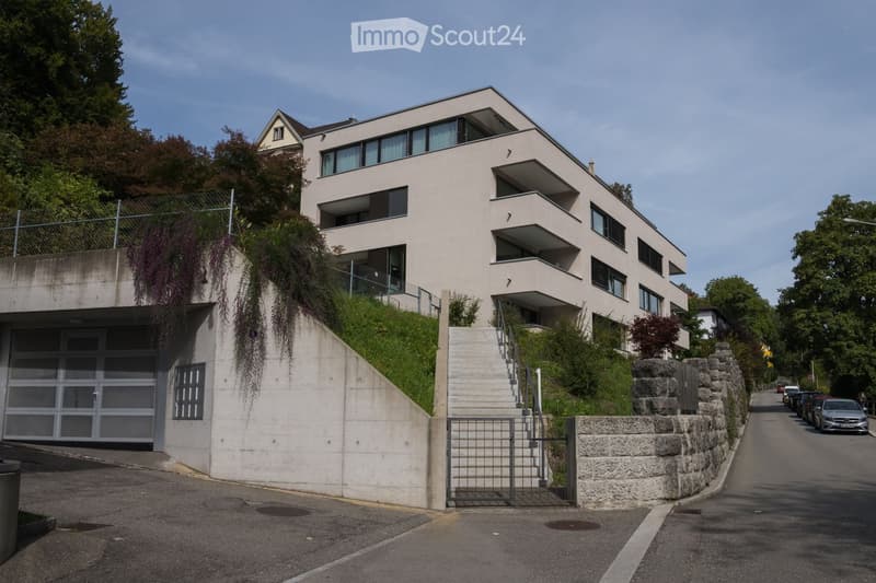 Modern multi-story building with large windows and balconies, situated on a raised ground floor with a concrete staircase leading up to the entrance.