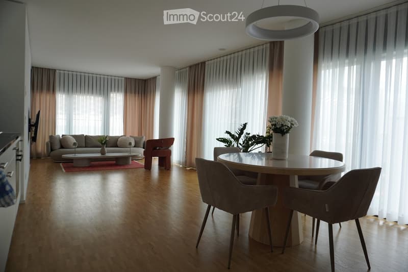 Open-plan living and dining area with wooden flooring, large windows with sheer curtains, a grey sofa, a round coffee table, a dining table with four chairs, and decorative vases with flowers.