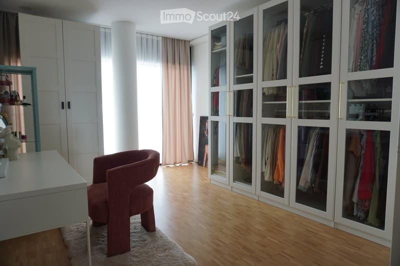 A well-lit room with wooden flooring, a pink chair, a white table, a large wardrobe with glass doors, and a mirror on the wall.