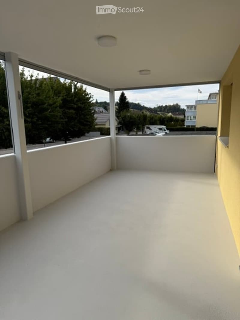 Empty balcony with white walls, large windows, and a view of the street and surrounding greenery.