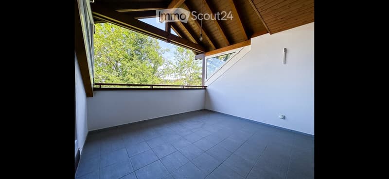 Empty room with wooden ceiling beams, large windows offering a view of greenery, tiled floor, and white walls.