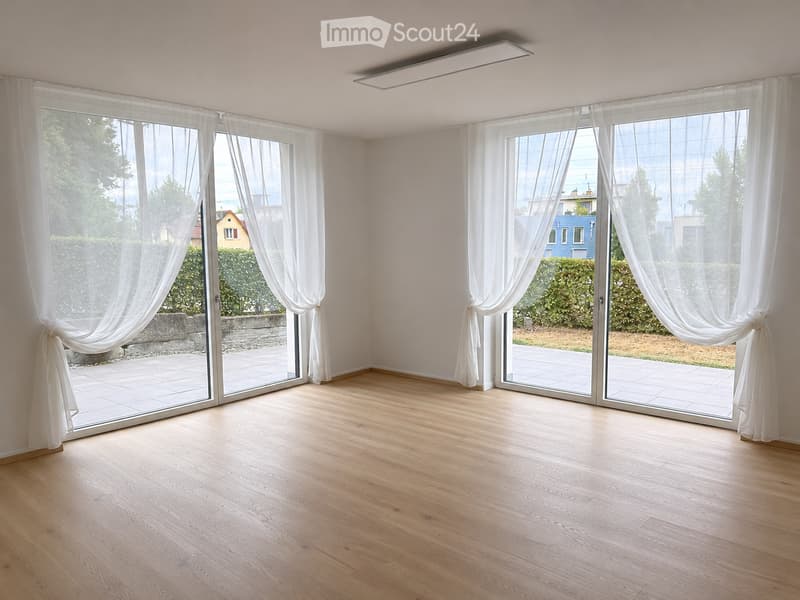 Empty room with wooden flooring, large sliding glass doors with white curtains, and bright natural light. No furniture or equipment present.