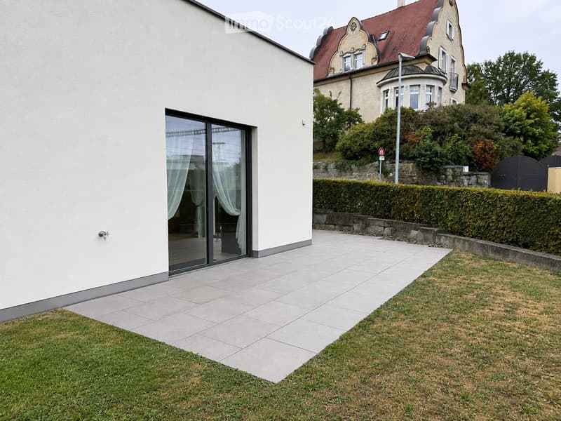 Garden area with a tiled patio, grassy lawn, and a hedge border. A modern house with large glass doors is visible on the left, and an ornate building is in the background.